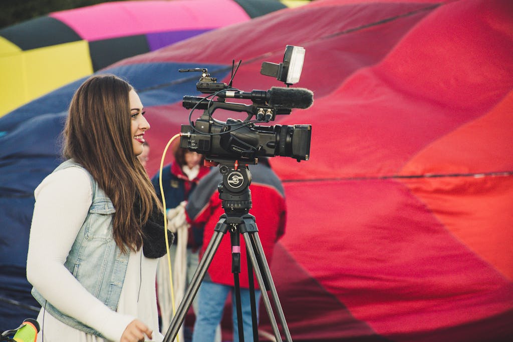 A female videographer operates a camera before a colorful hot air balloon backdrop.