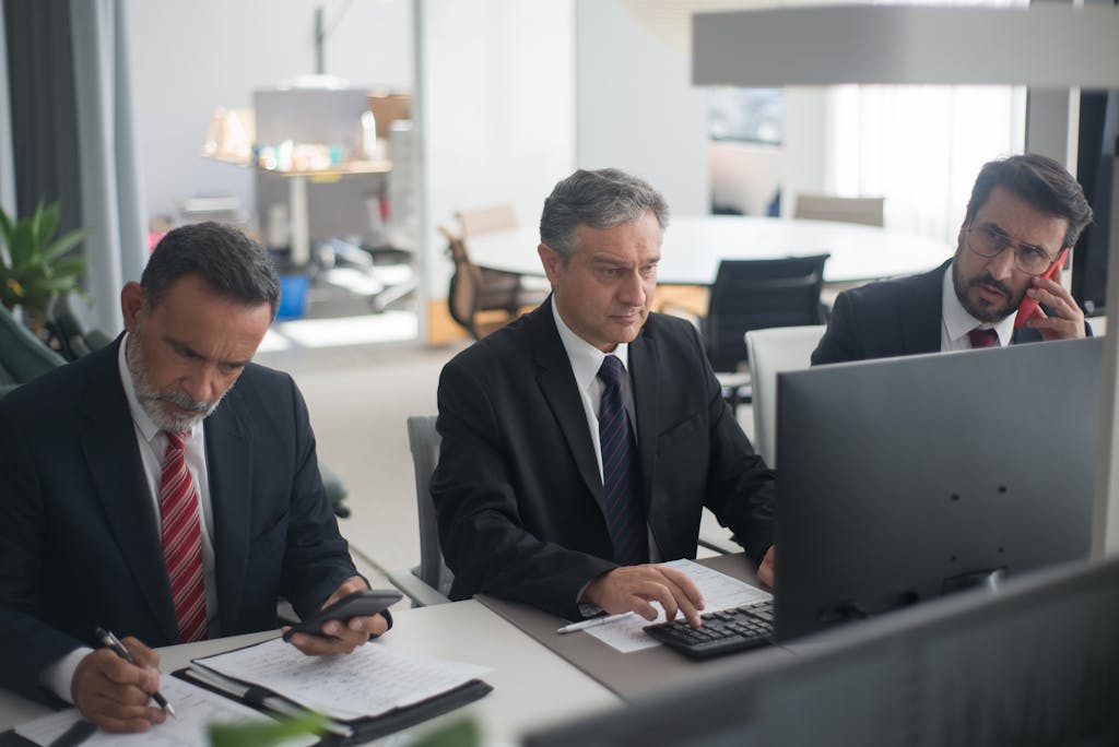 Three businessmen in suits working on computers and documents in a modern office setting.